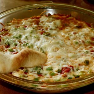 Pepperoni pizza dip in a round glass pie plate with mozzarella, green pepper and green onion on top and a soft breadstick dipped in