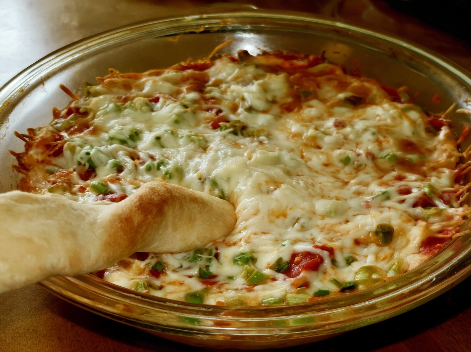 Pepperoni pizza dip in a round glass pie plate with mozzarella, green pepper and green onion on top and a soft breadstick dipped in