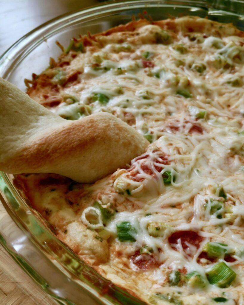 Close-up of pepperoni pizza dip in a glass pie plate with two soft breadsticks dipped in, showing green pepper, green onion and bits of chopped pepperoni under the mozzarella