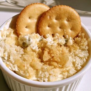 Jalapeño popper dip in a white ramekin with a crumbly toasted parmesan top and two round butter crackers standing in the dip