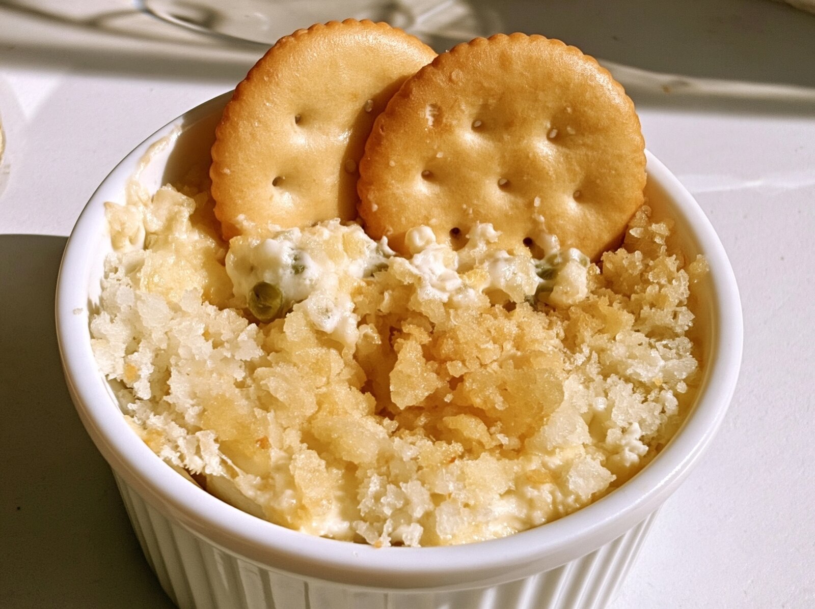 Jalapeño popper dip in a white ramekin with a crumbly toasted parmesan top and two round butter crackers standing in the dip