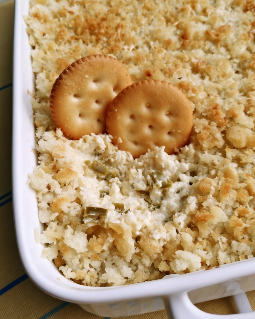 Jalapeño popper dip in a white square baking dish with a toasted parmesan crumb top, two round butter crackers standing in a scooped corner showing diced green peppers in the creamy dip