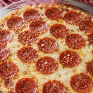 Pepperoni pizza dip fresh from the oven in a glass pie dish with bubbling mozzarella and crispy pepperoni slices on a white surface with a red gingham towel in the background.