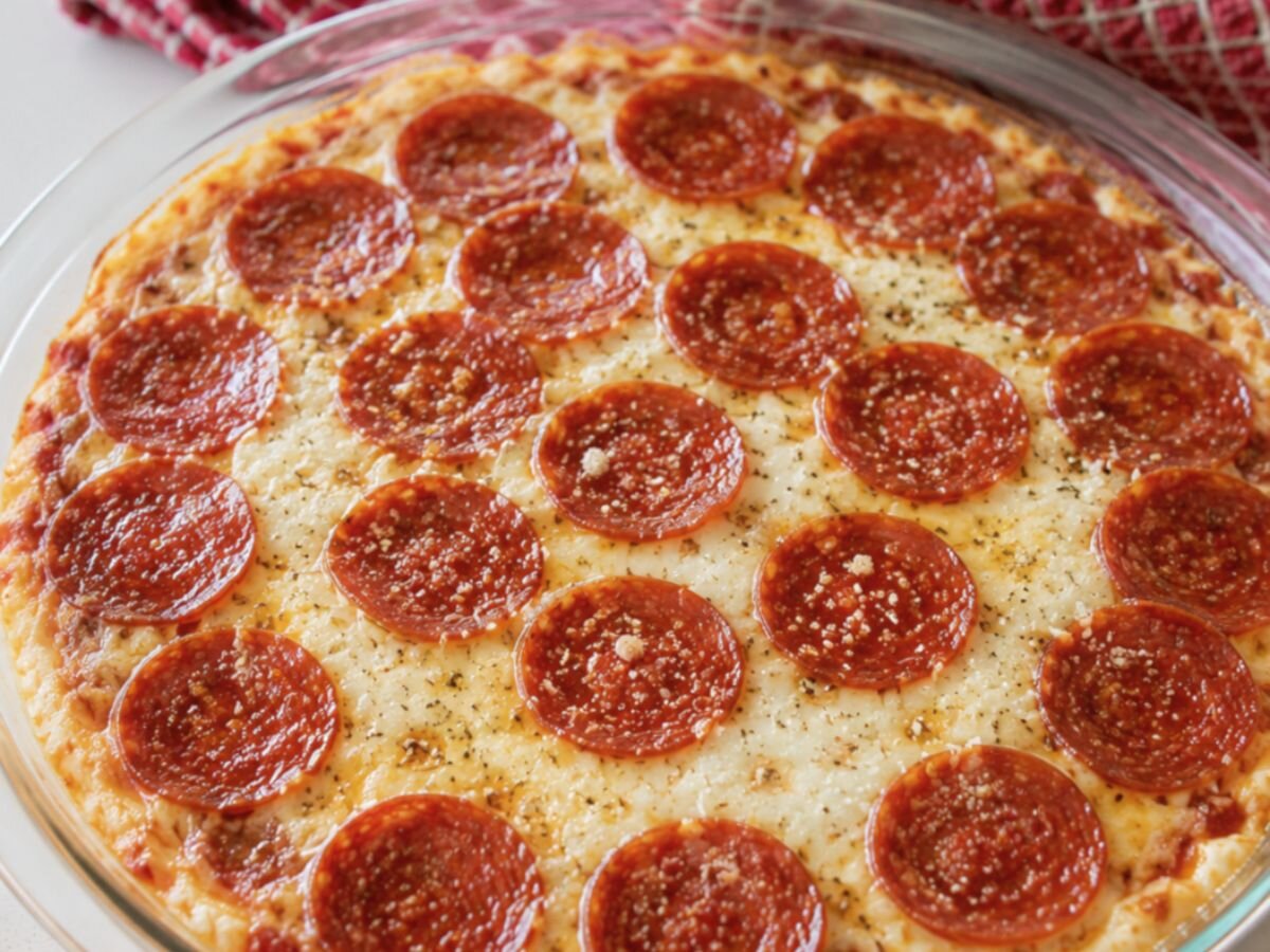 Pepperoni pizza dip fresh from the oven in a glass pie dish with bubbling mozzarella and crispy pepperoni slices on a white surface with a red gingham towel in the background.