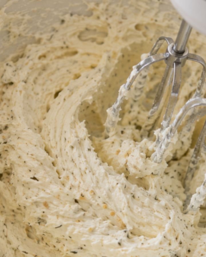 Hand mixer beating softened cream cheese with garlic powder and Italian seasoning into a smooth fluffy mixture in a white bowl on a white quartz countertop.