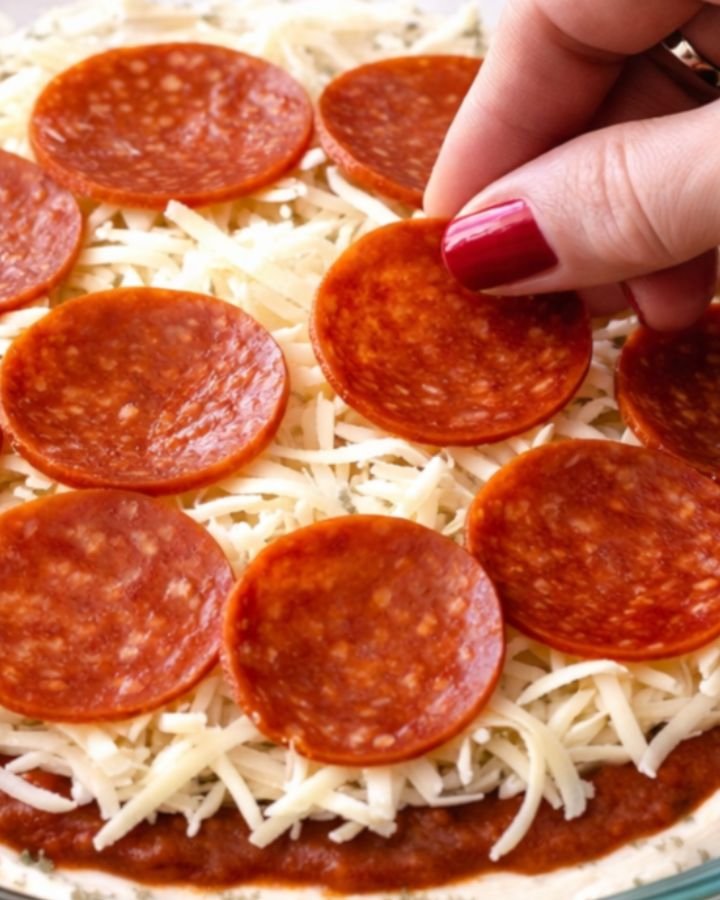 Hand placing pepperoni slices over shredded mozzarella cheese in a glass pie dish showing the assembled layers of pizza dip before baking.